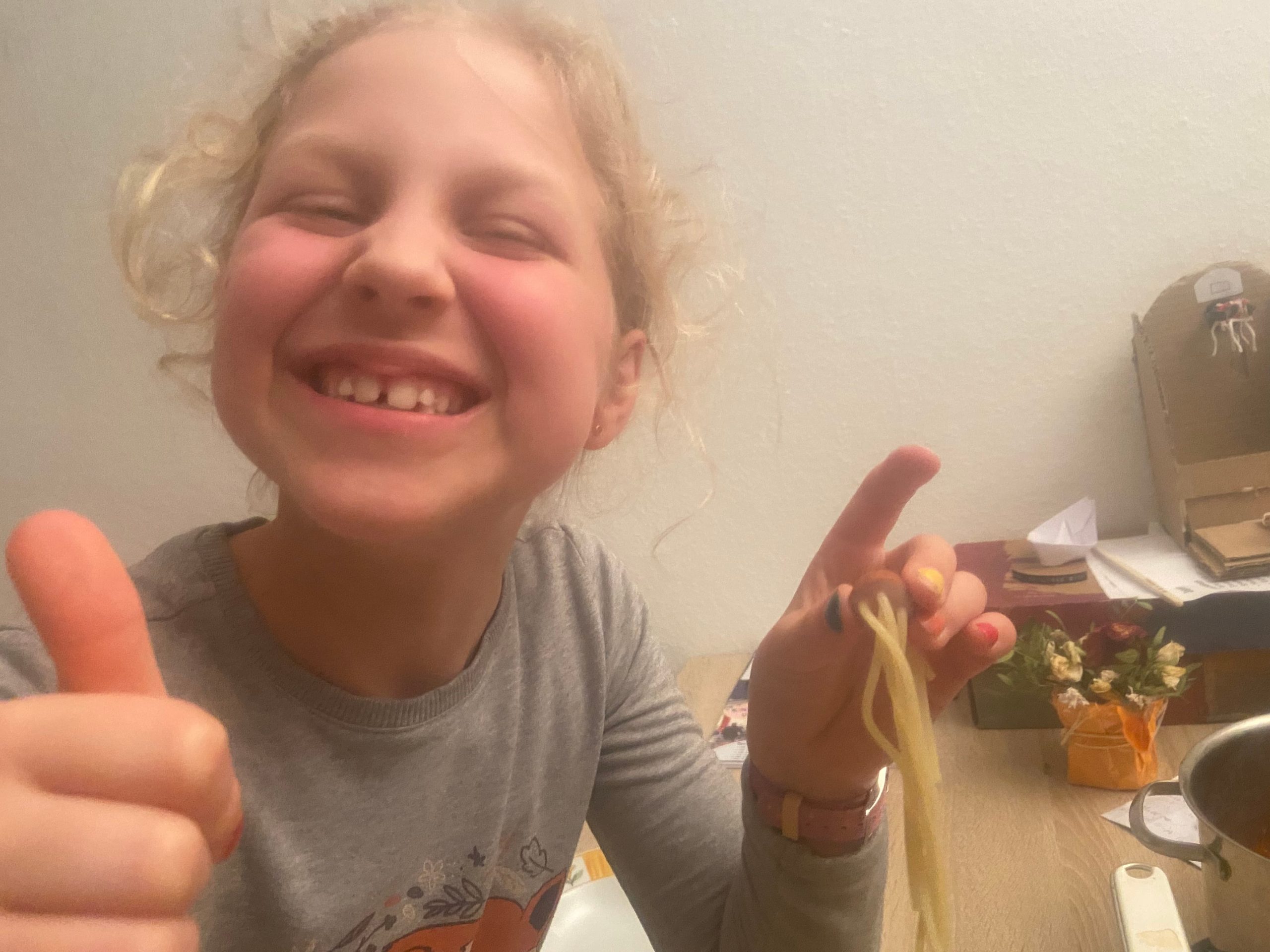 Elsina-Sophie smiling, giving thumbs-up and holding spaghetti strung through a hotdog